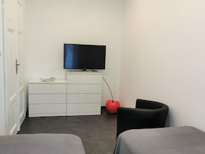 Une petite pièce moderne avec un téléviseur à écran plat fixé sur un meuble blanc, un ballon rouge pour faire de l’exercice, deux chaises noires et un canapé gris.