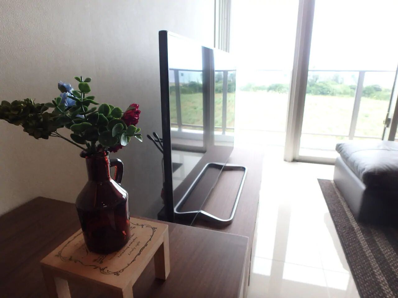 Uma pequena mesa de madeira com um vaso escuro contendo uma planta com flores azuis e vermelhas, um dispositivo eletrônico preto e uma bandeja de metal.