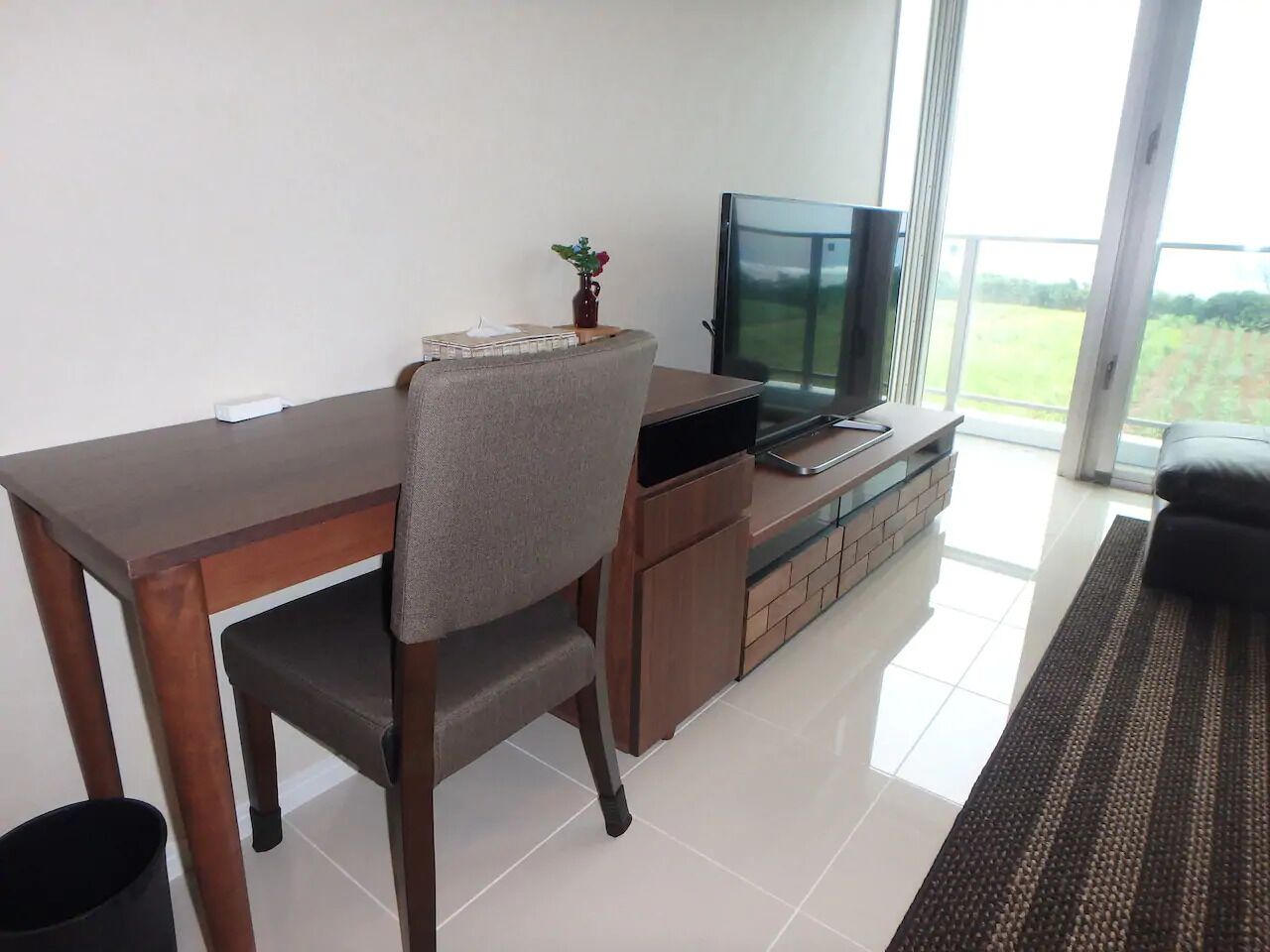 A modern living room with a wooden dining table, a grey upholstered chair, a TV on a wooden cabinet, and a view of a green landscape through large windows.