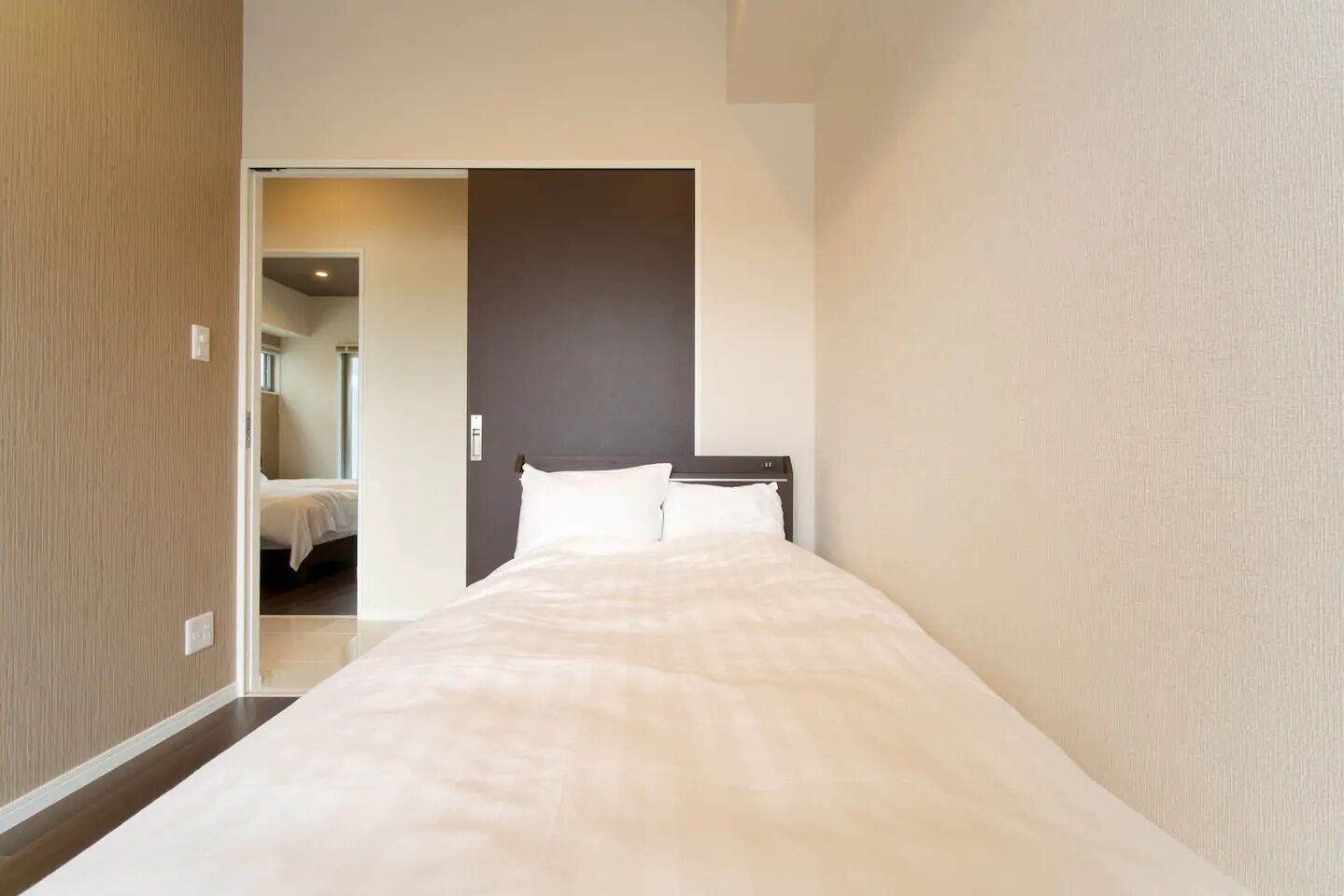 A narrow hotel room with a bed, a dark headboard, and a door leading to another room.