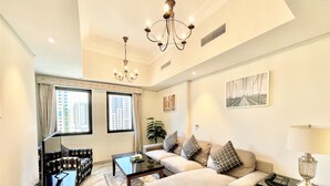 A modern living room with a sofa, coffee table, and a chandelier. There are two windows with curtains, a plant, and artwork on the walls. - Spacious Marbella 2BR With Scenic Views (Dubai)