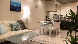 A modern living room with a glass coffee table, a white sofa with patterned cushions, a dining area with a glass table, and a kitchen in the background. - Longvacation Okinawa 206 (Onna)