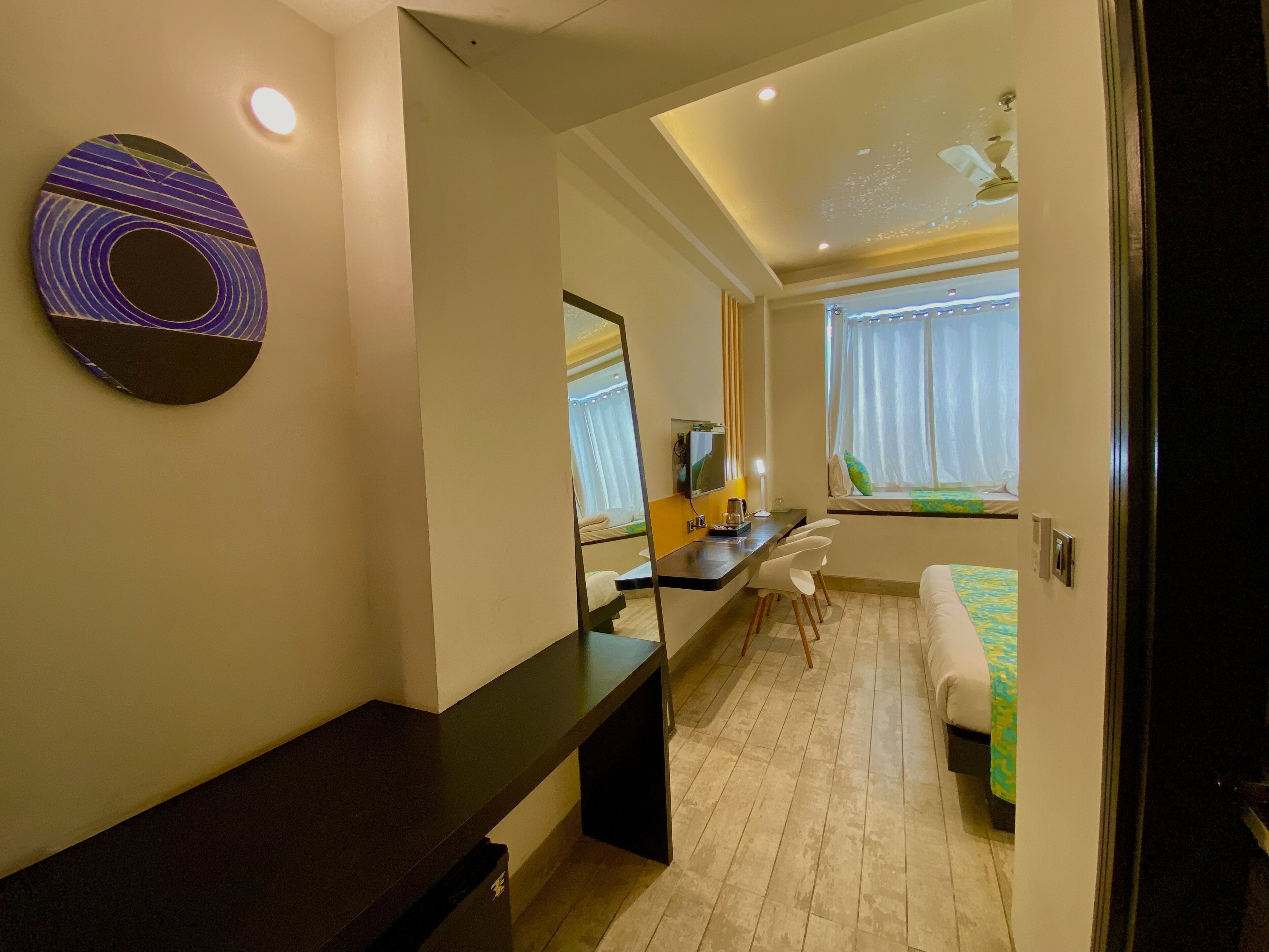 A modern hotel room with a desk, a bed, a large mirror, and a window with curtains.
