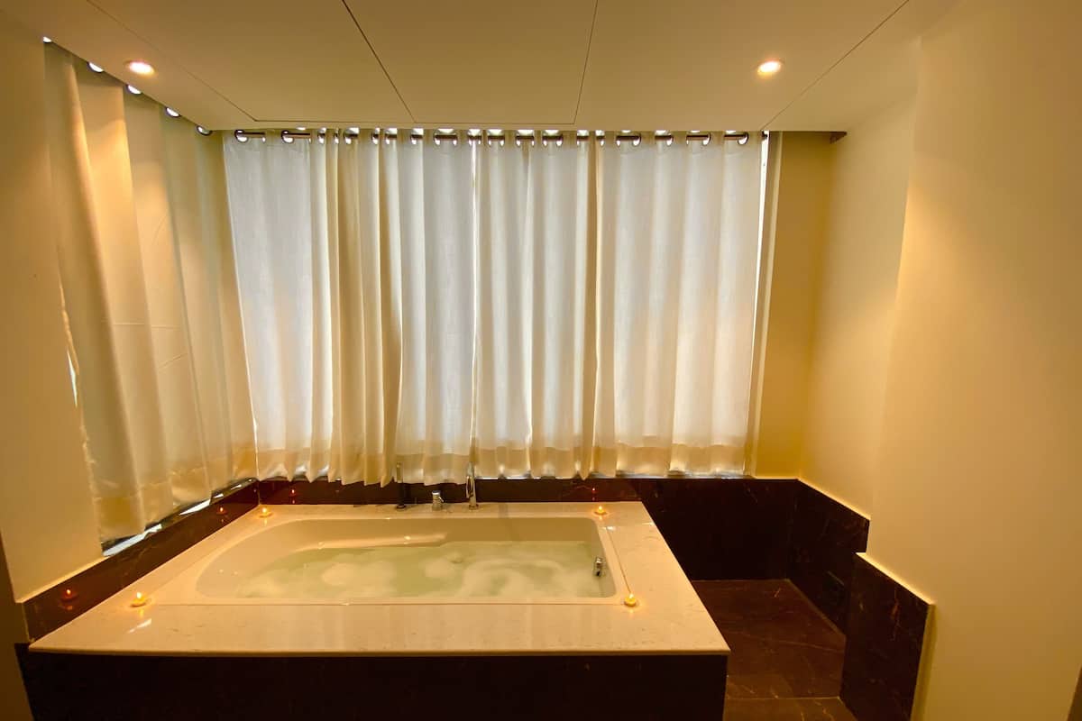 A large rectangular bathtub with a faucet, surrounded by dark marble flooring, and a window with sheer curtains.