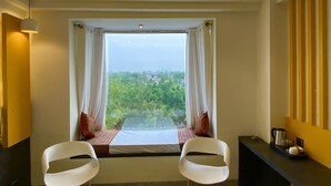 Una habitación con una cama, dos sillas blancas, un escritorio y vistas al exterior con vegetación.