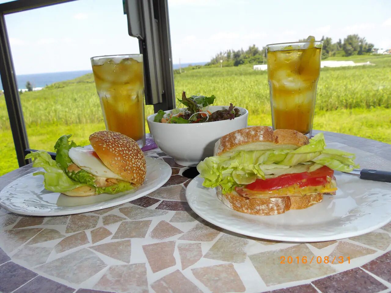 Een tafel met twee sandwiches, een salade en twee glazen ijsthee, met uitzicht op een groen veld en de zee.
