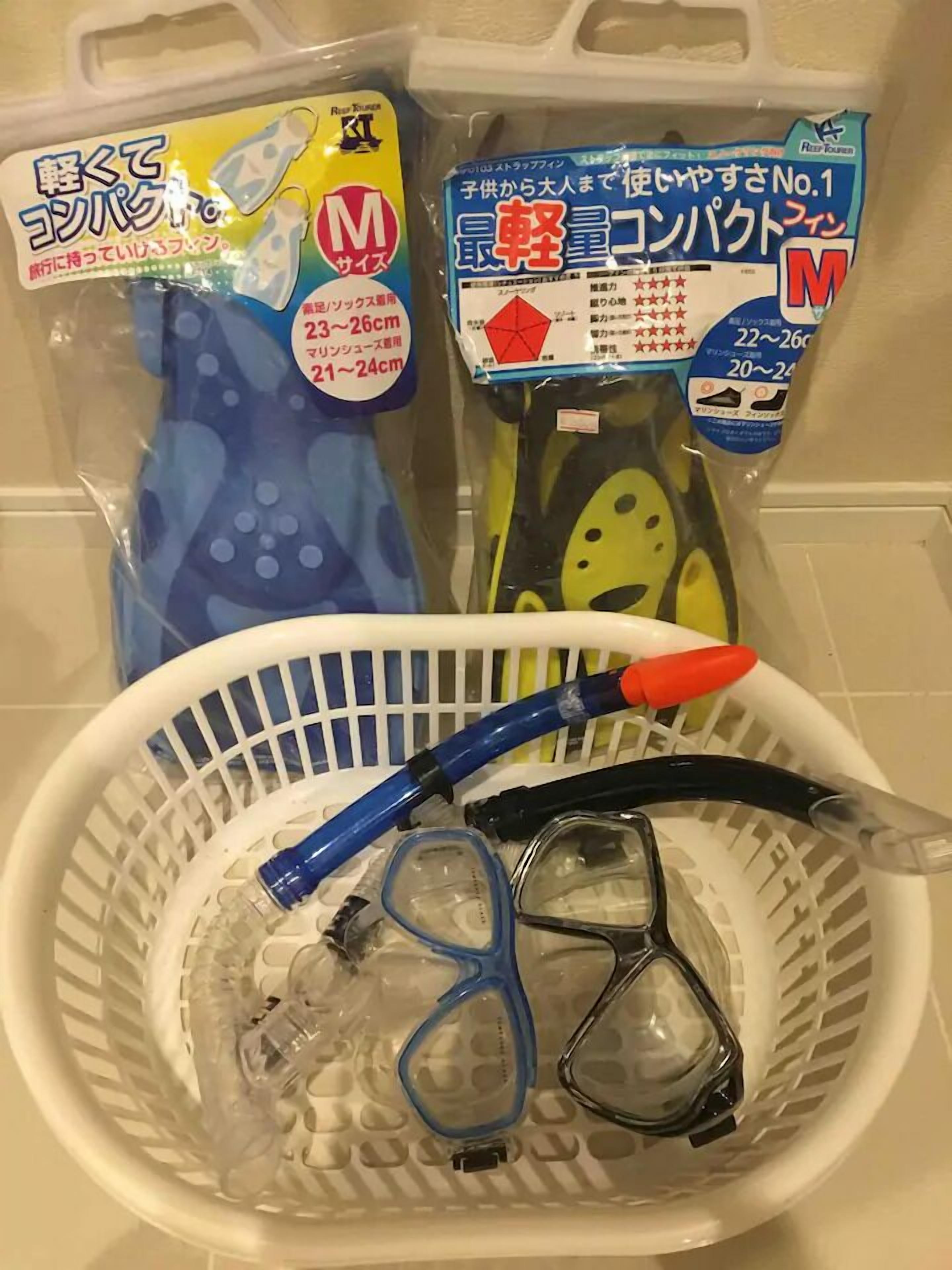 A white plastic basket containing a blue snorkel, a pair of black and clear diving goggles, and a yellow and blue swim mask.