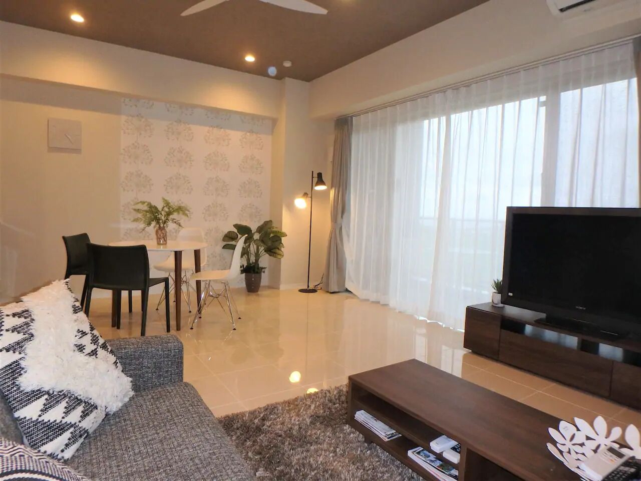 A modern living room with a flat-screen TV, a grey sofa with patterned cushions, a coffee table, and a dining area with chairs and a table.