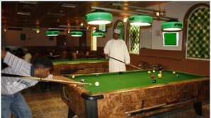 A pool table in a room with green hanging lights, a patterned wall, and a person playing pool. - Bowshar International Hotel (Muscat)