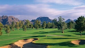 Um campo de golfe com bunkers, árvores e montanhas ao fundo.