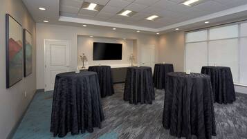 A conference room with a large flat-screen TV, multiple round tables covered with black tablecloths, and a carpeted floor.
