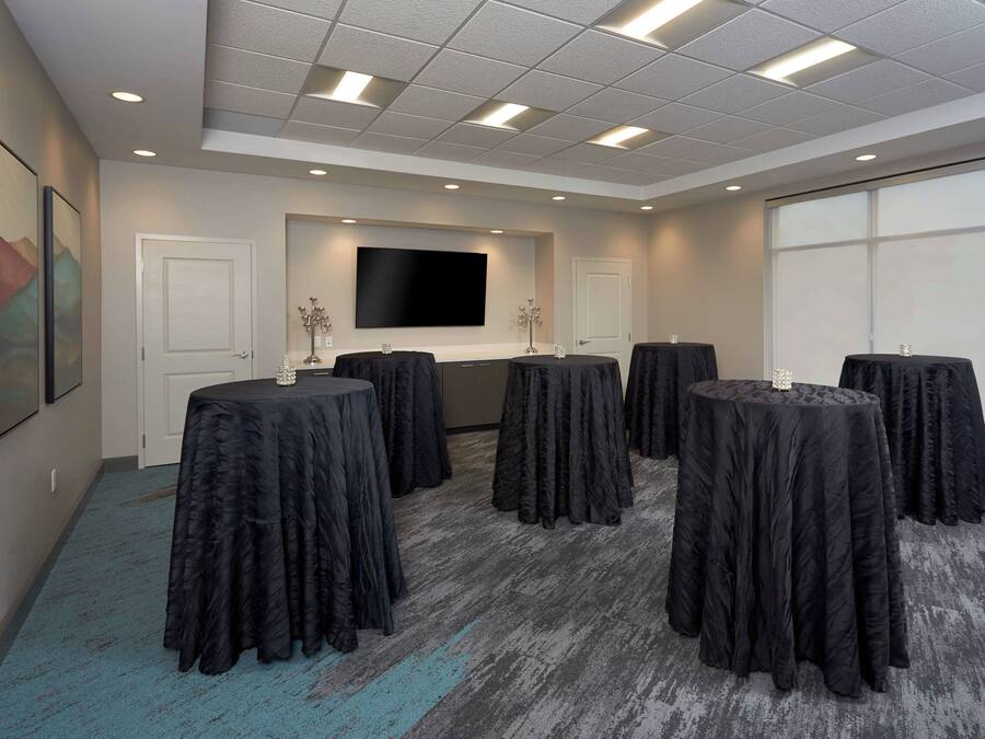 A conference room with a large flat-screen TV, multiple round tables covered with black tablecloths, and a carpeted floor.