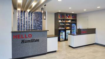 Uma área de recepção com um balcão identificado como "HELLO Hamilton", parede com padrão, máquina de vendas automática e um refrigerador.