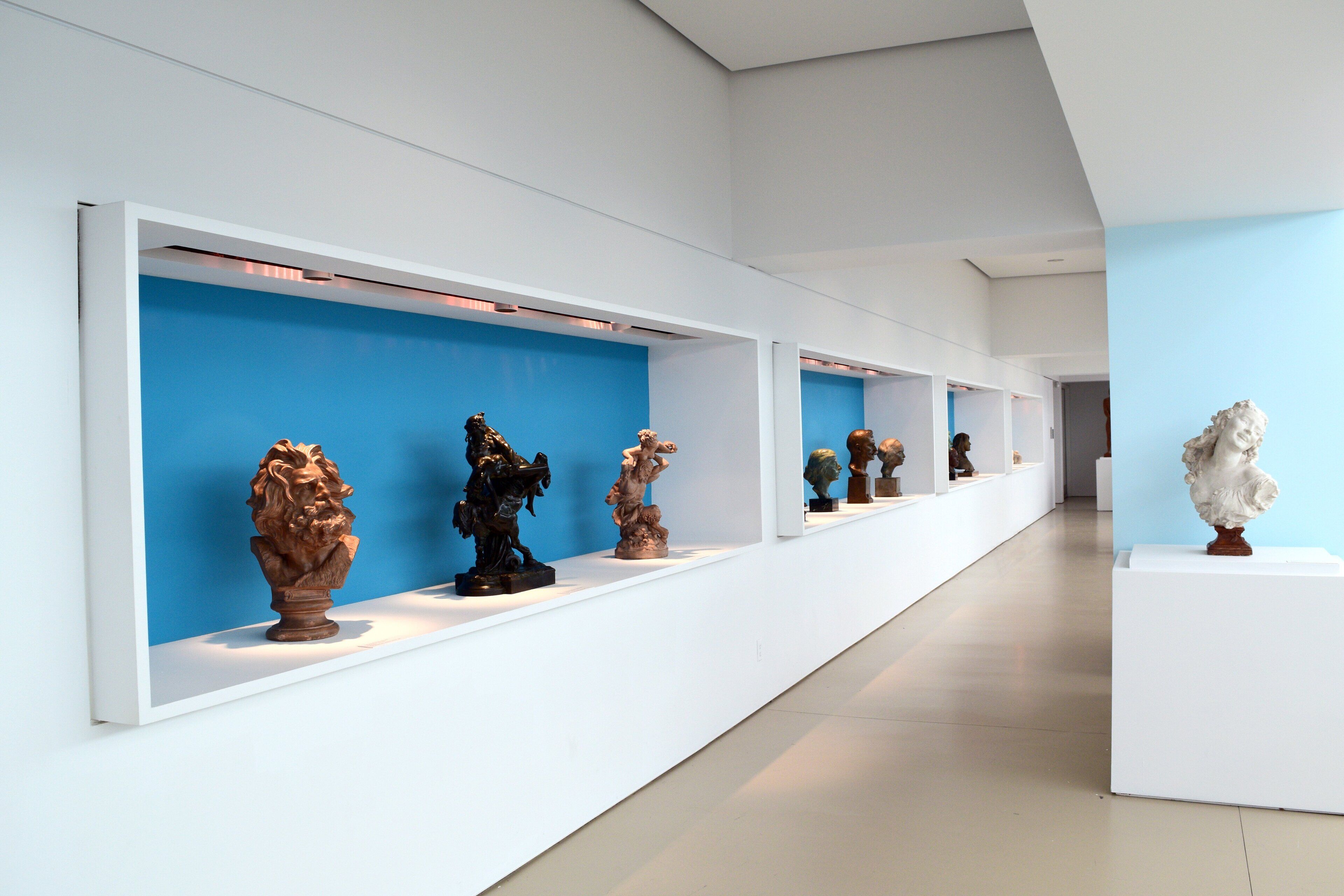 Et moderne kunstgalleri med hvide vægge og blå baggrunde, der udstiller forskellige skulpturer på hylder.