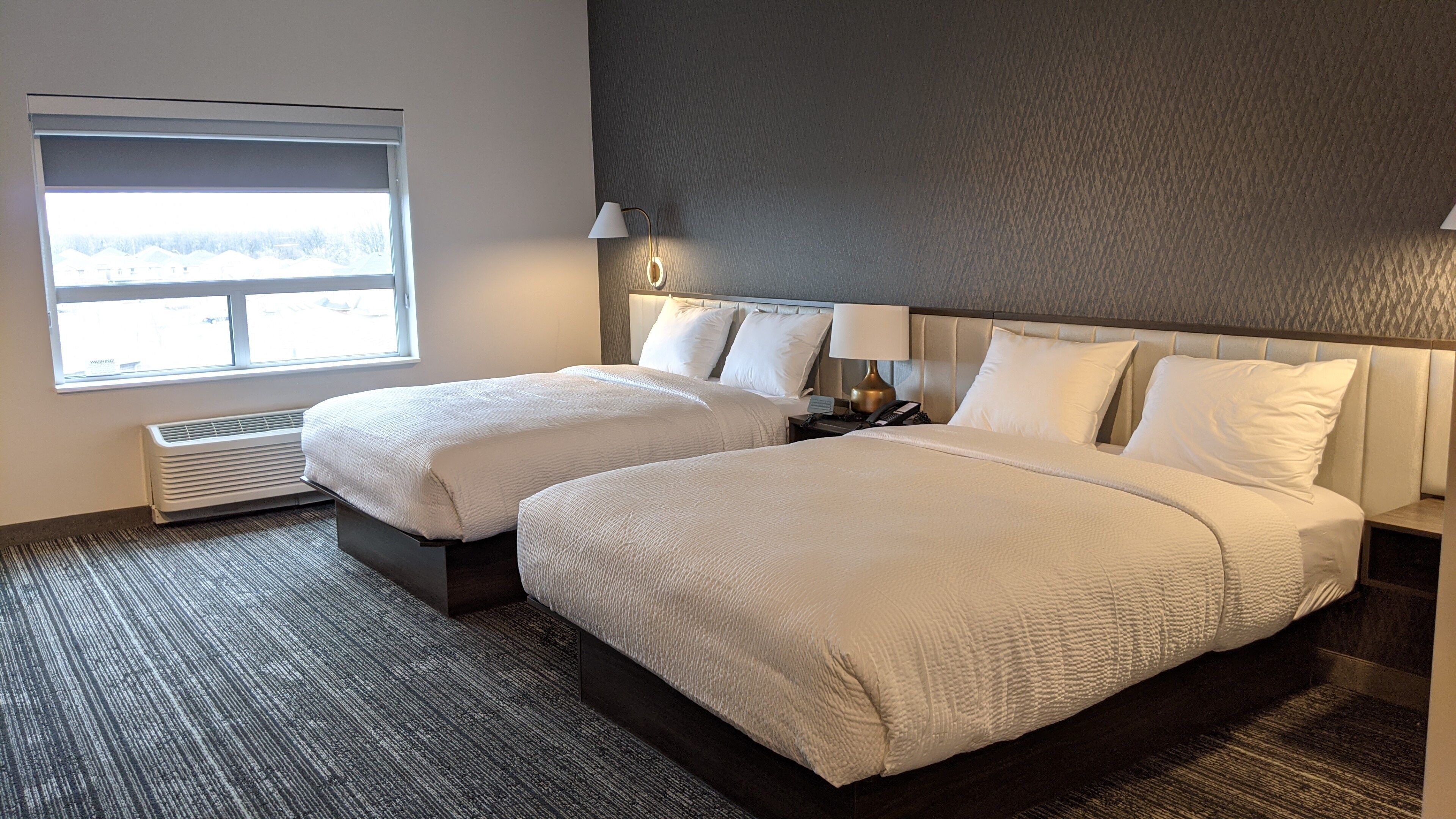 A hotel room with two beds, a large window, a wall heater, and a nightstand with a lamp.
