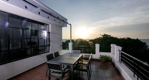 A rooftop terrace with a table and chairs, a large window, and a view of the sunset over the sea. - Pikidi Gardens (Bondo)