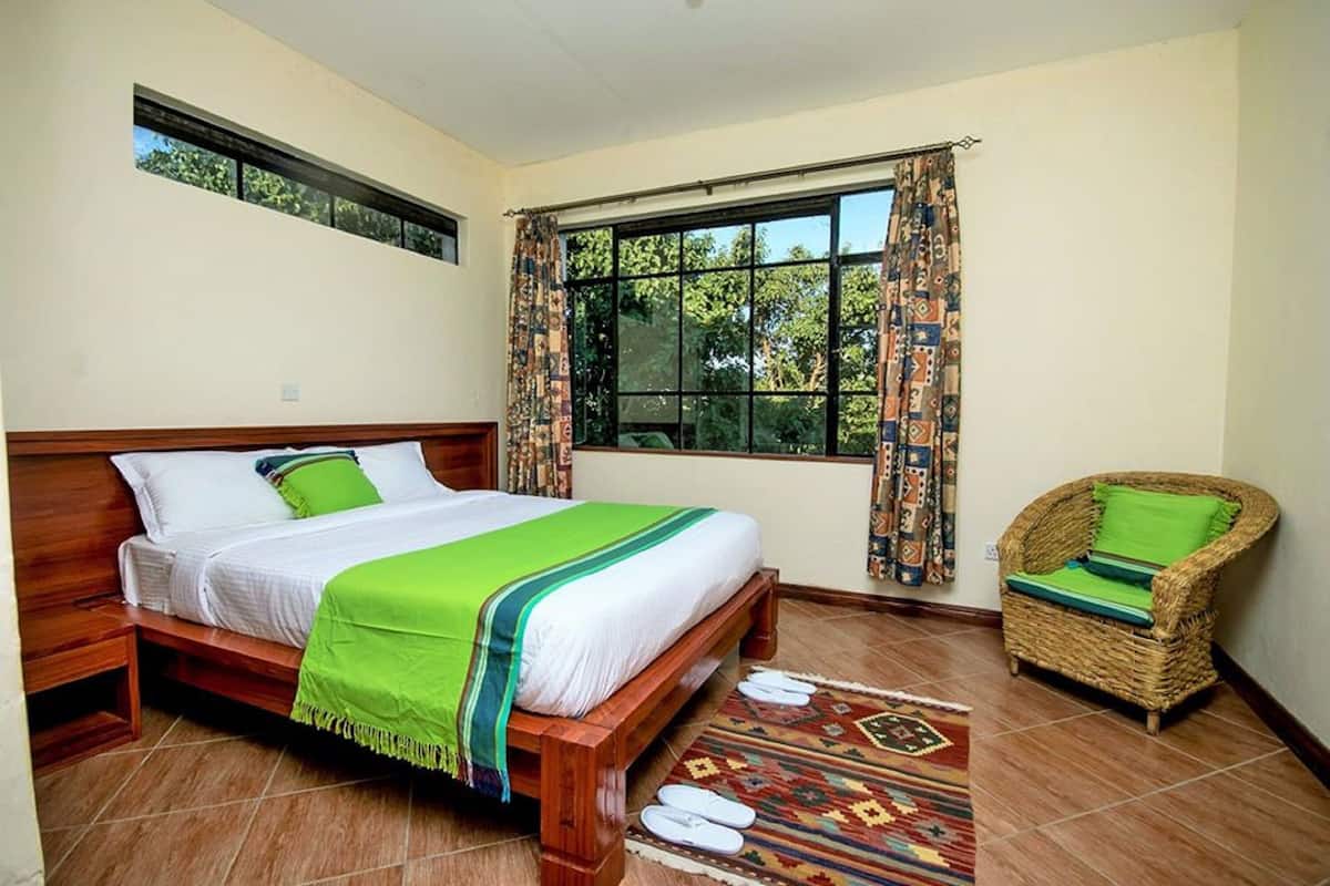 Quarto com cama de madeira, colcha verde e branca, cadeira de vime com almofada verde, janela grande com cortinas estampadas e uma pequena janela acima da cama.