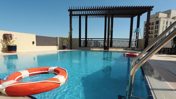 A swimming pool with a lifebuoy, a covered seating area, and a view of buildings in the background. - Marbella Executive 2 Bedrooms (Dubai)