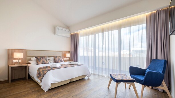 A hotel room with a bed, bedside tables, a chair, a small table, and a large window with curtains. - Casa Amarela 22 (Azores Premium at city center) (Ponta Delgada)