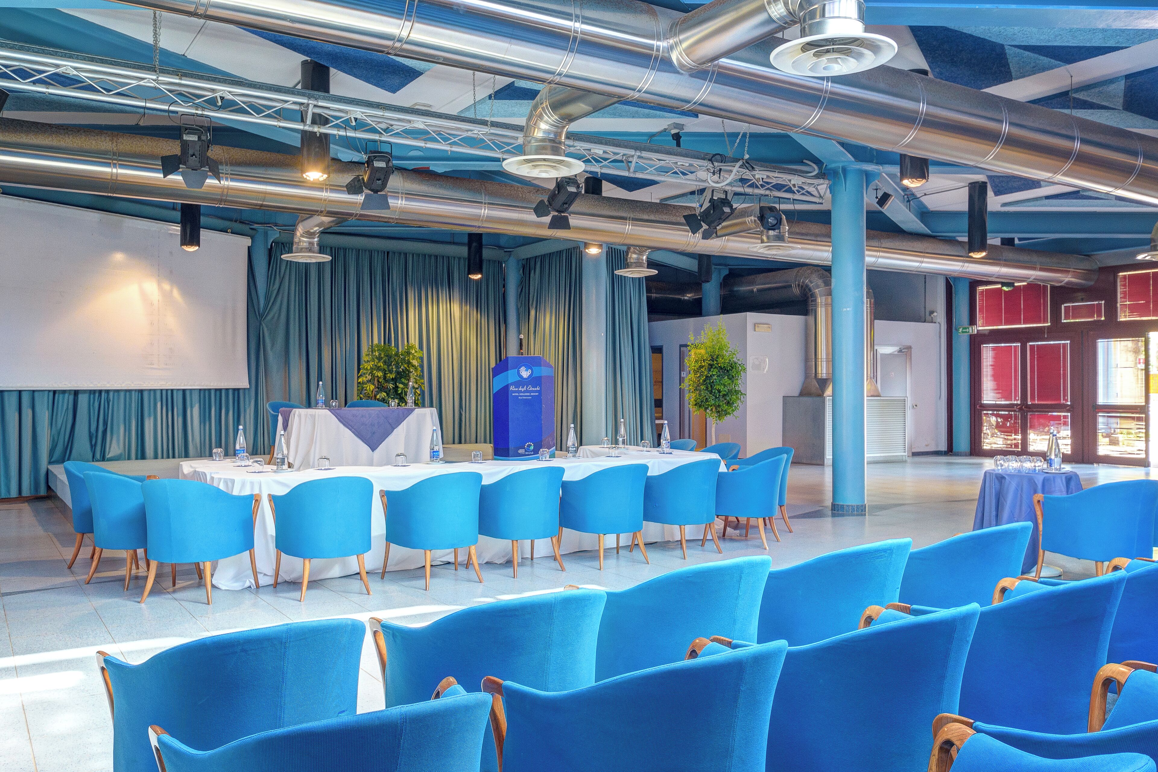 A conference room with blue chairs and a white table, large windows, and a stage with curtains.