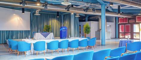 A conference room with blue chairs and a white table, large windows, and a stage with curtains.