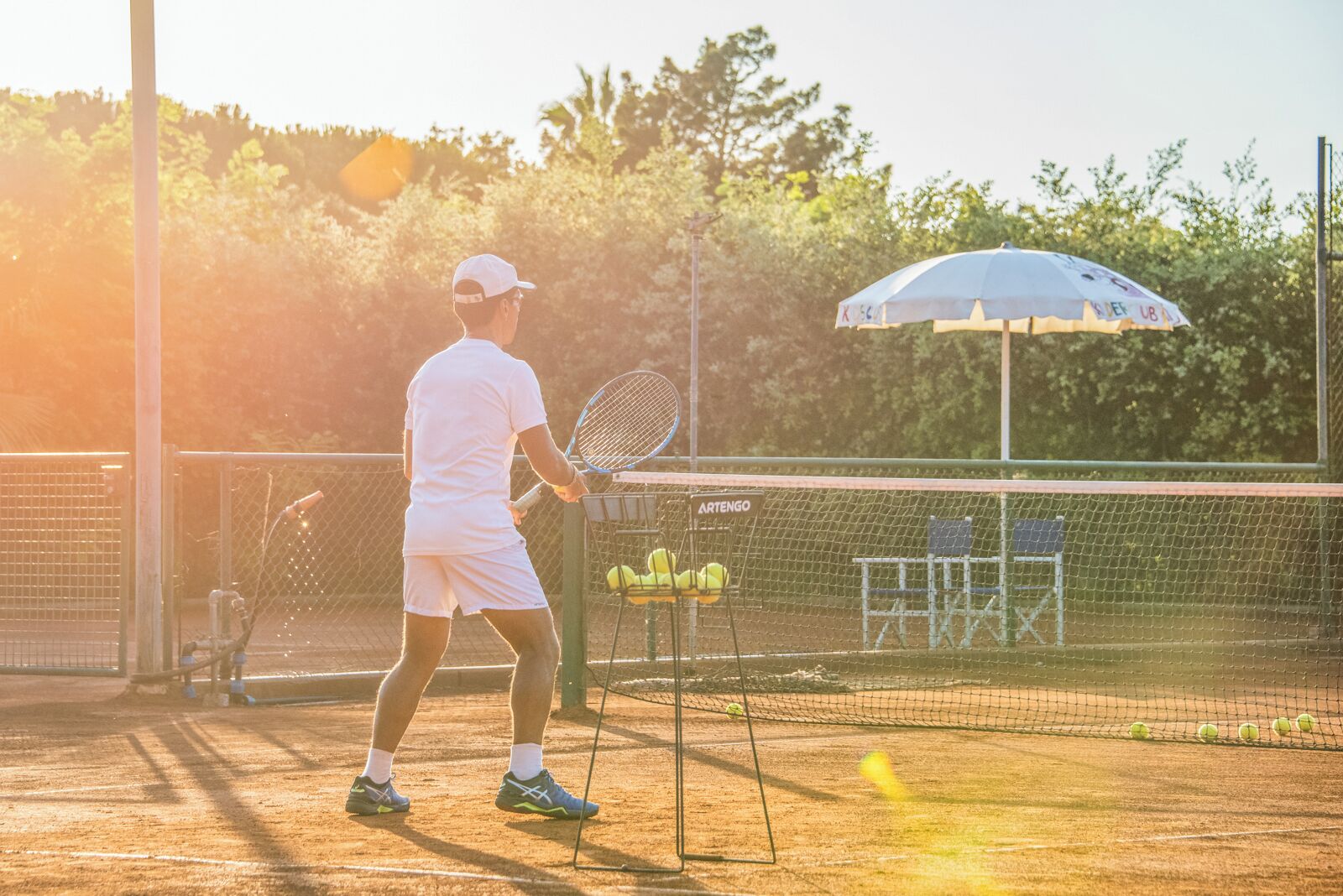 Un campo da tennis con un giocatore, una rete e una panchina piena di palline da tennis.