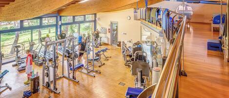 A gym with wooden beams, exercise equipment, and large windows overlooking a green area.