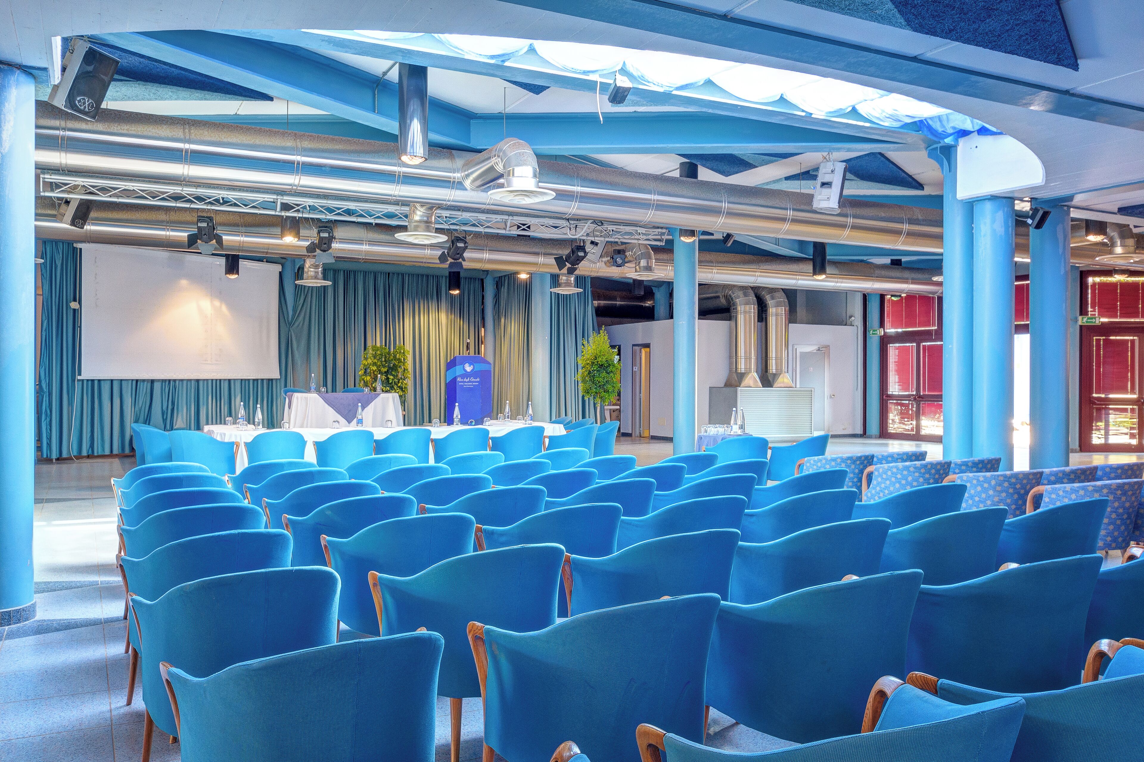 Un salón de conferencias con filas de sillas azules, una pantalla grande y un escenario con cortinas.