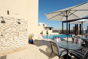 A rooftop pool area with a glass table and chairs, a pergola with a large umbrella, and a view of the city skyline.