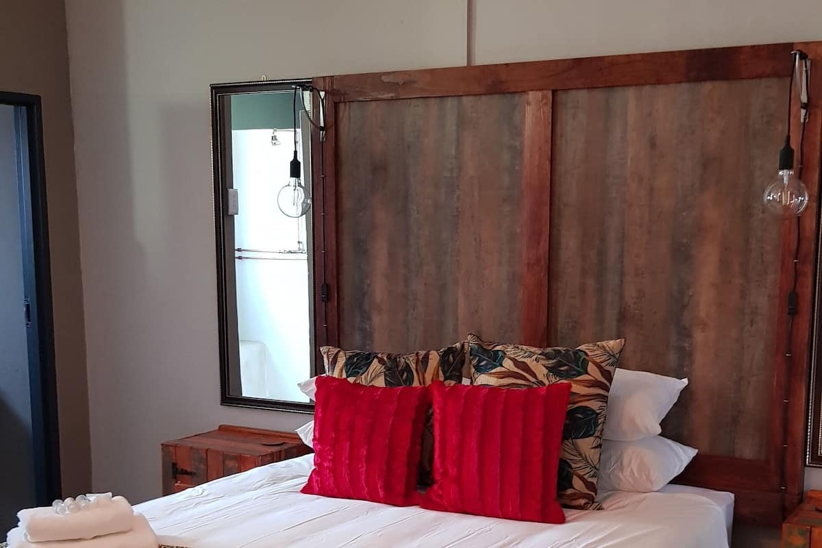 A neatly made bed with white linens, red pillows, and a tropical leaf-patterned pillow. A wooden headboard and a hanging light fixture are visible.