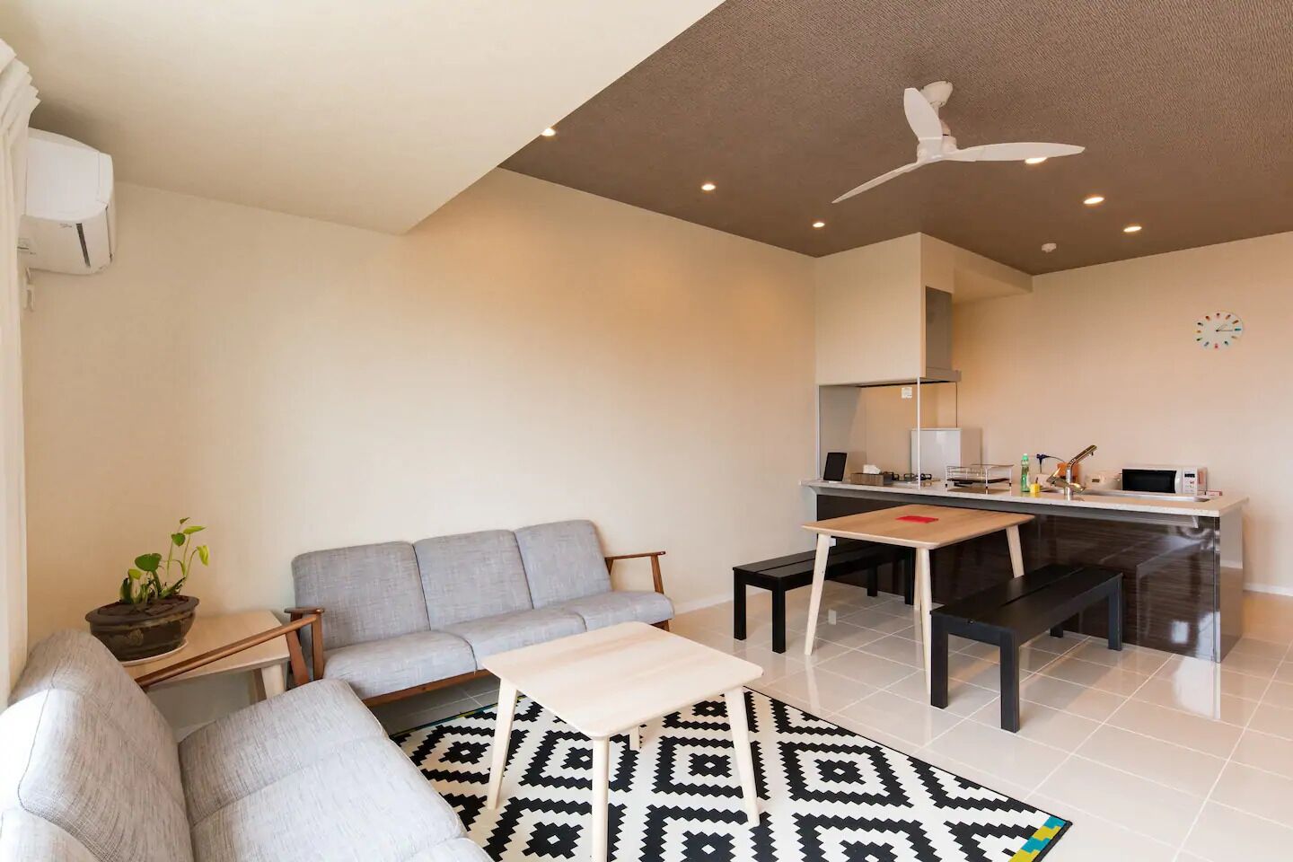 Una sala de estar moderna con un sofá gris, una mesa de centro y un tapete estampado. Hay una mesita auxiliar con una planta. La habitación tiene un ventilador de techo e iluminación empotrada.