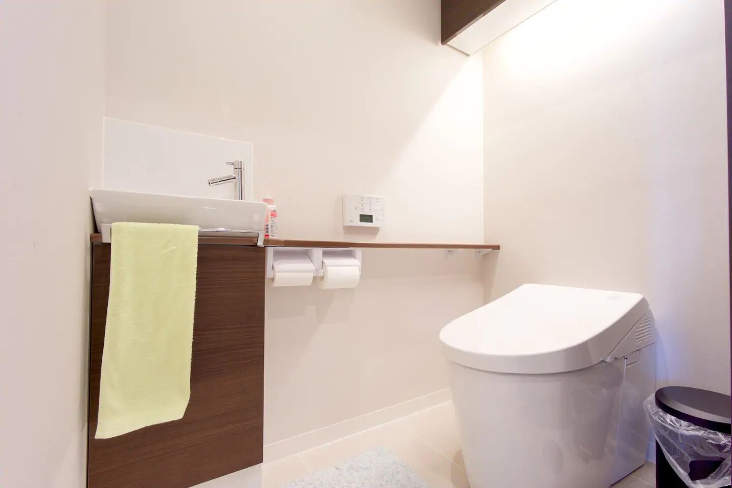 Un baño moderno con inodoro, una toalla colgada en un panel de madera, estante con artículos de tocador y un panel de control en la pared.