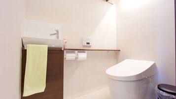 Un baño moderno con inodoro, una toalla colgada en un panel de madera, estante con artículos de tocador y un panel de control en la pared.