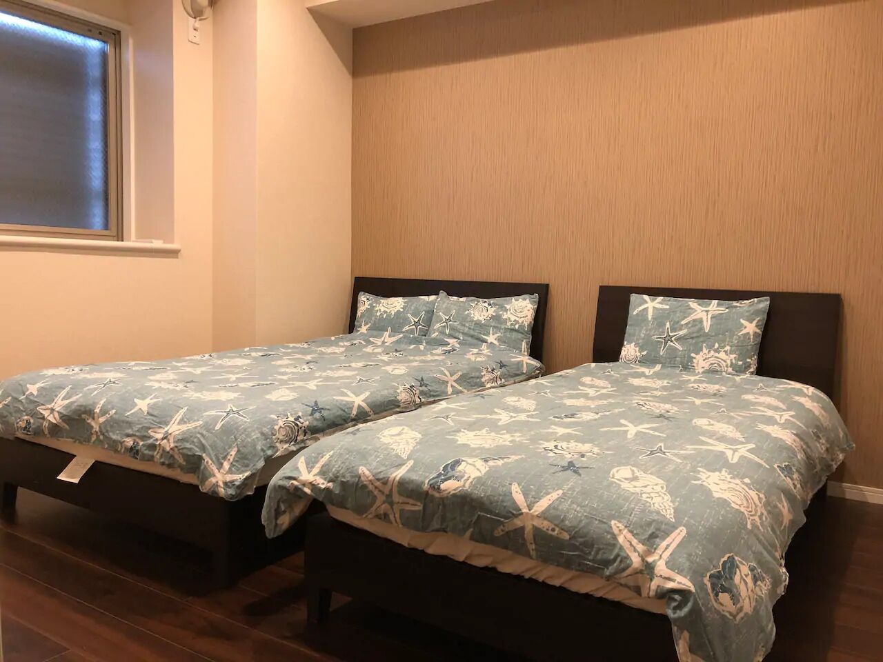 Two twin beds with matching blue bedding featuring sea creatures, positioned against a wooden wall with a window above.