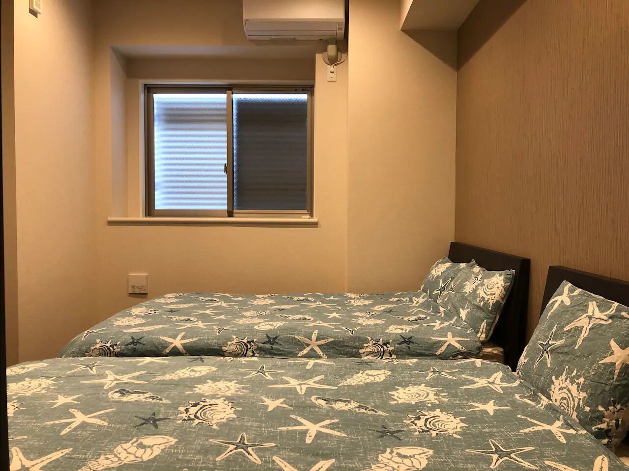 A bedroom with a bed featuring a nautical-themed bedspread, a window with blinds, and a wall-mounted air conditioning unit.