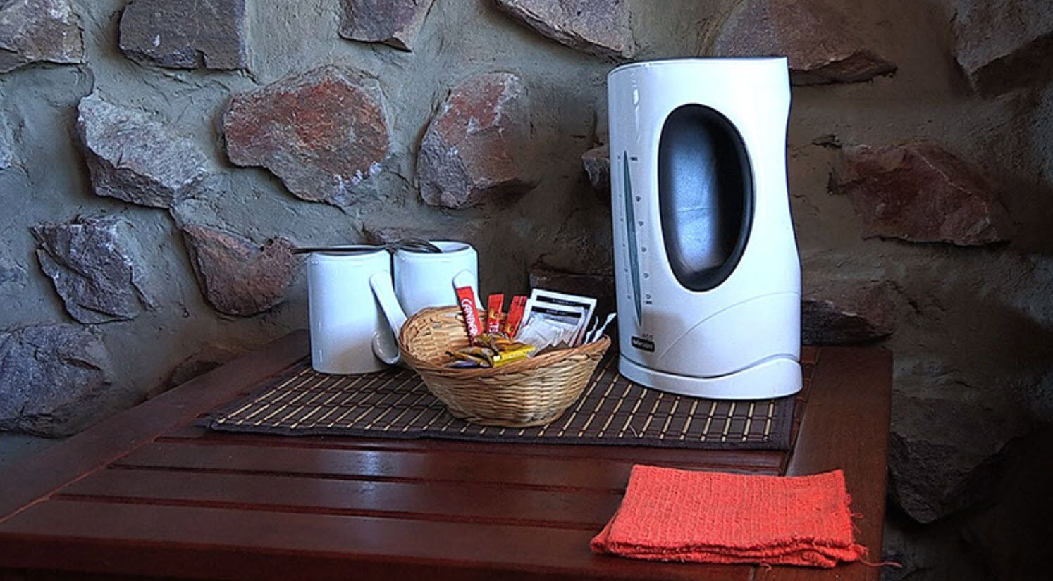 A stone wall, a white kettle, a basket with snacks, and a red napkin on a wooden table.