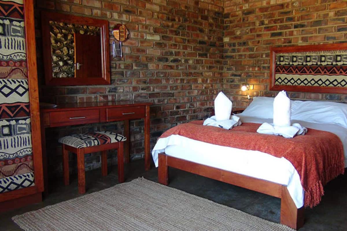 A room with a brick wall, a bed with white linens, a wooden desk, a chair, and a wall-mounted mirror.