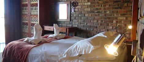 Habitación de hotel con pared de ladrillo, cama con ropa blanca, manta roja y ventana con luz natural.