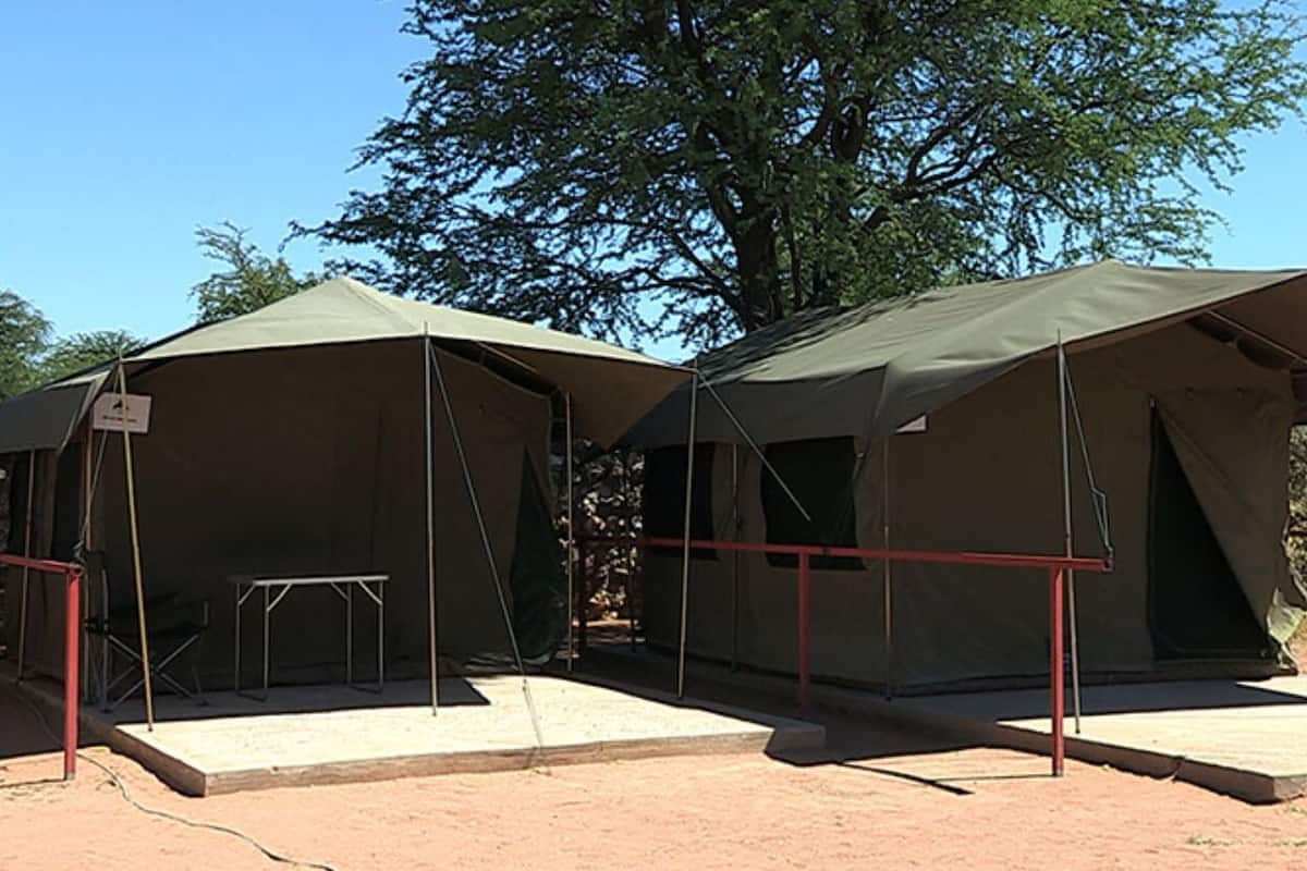 Three tents with a table and chairs in front, set up in a dry, open area with trees in the background.