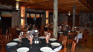 A dining area with round tables set for a meal, wooden chairs, and a brick wall in the background. - West Nest Lodge (Witvlei)
