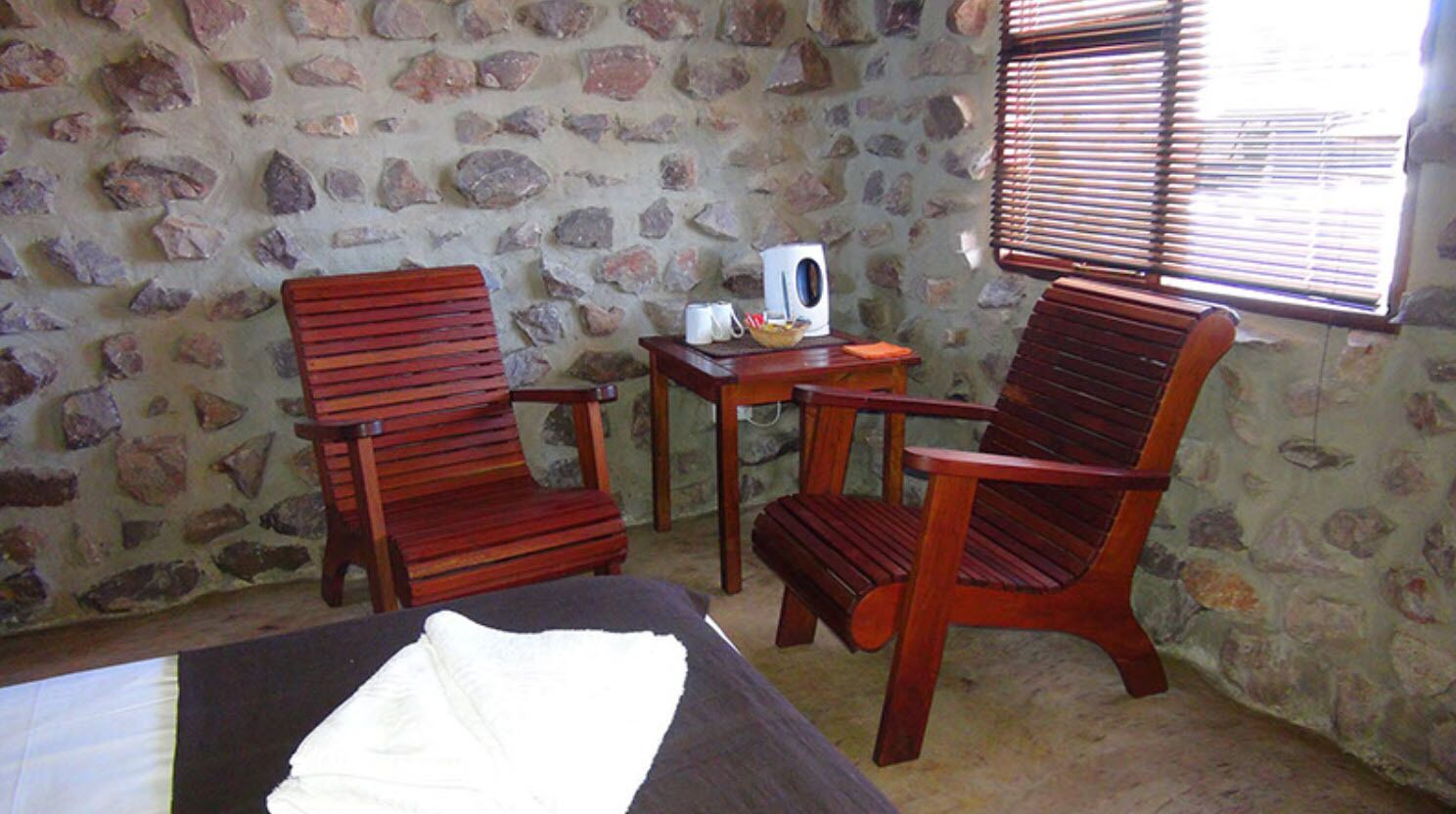 A room with a stone wall, a bed, two wooden chairs, a small table with a kettle and a bowl, and a window with blinds.