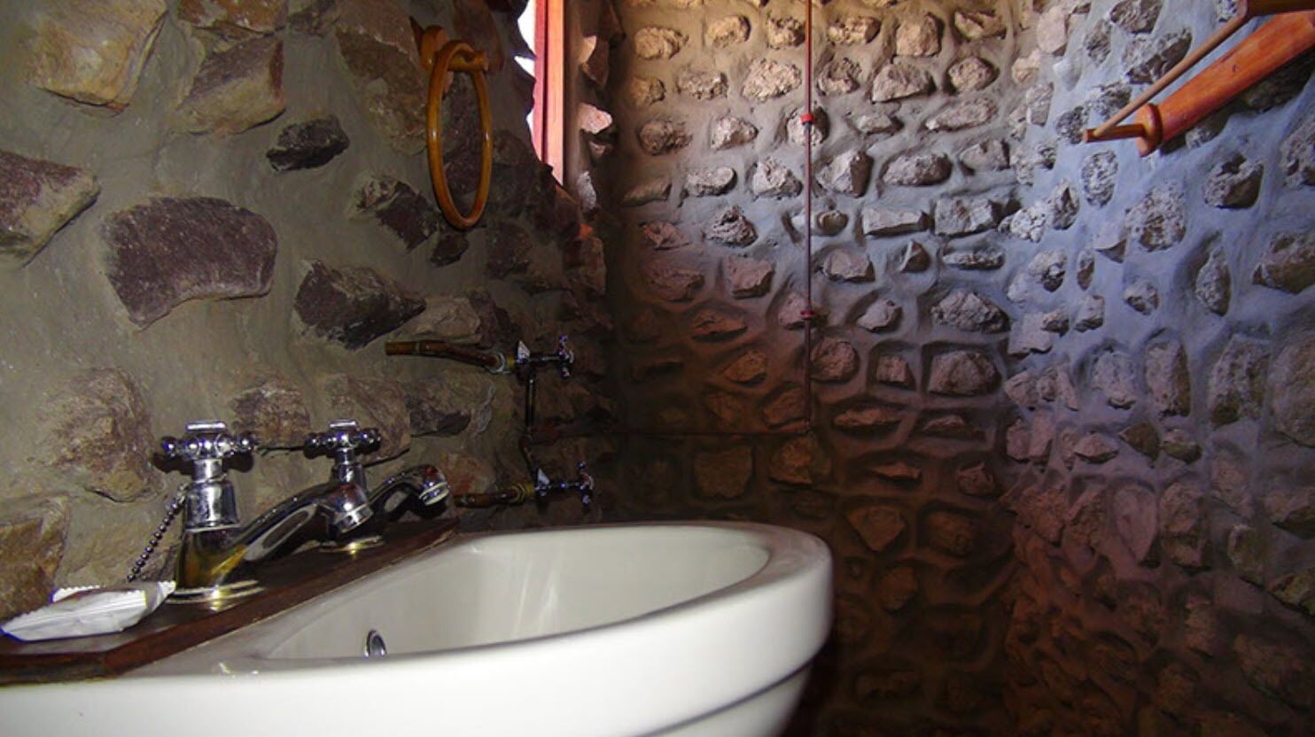A stone-walled bathroom with a white sink, two faucets, and a wooden shelf.