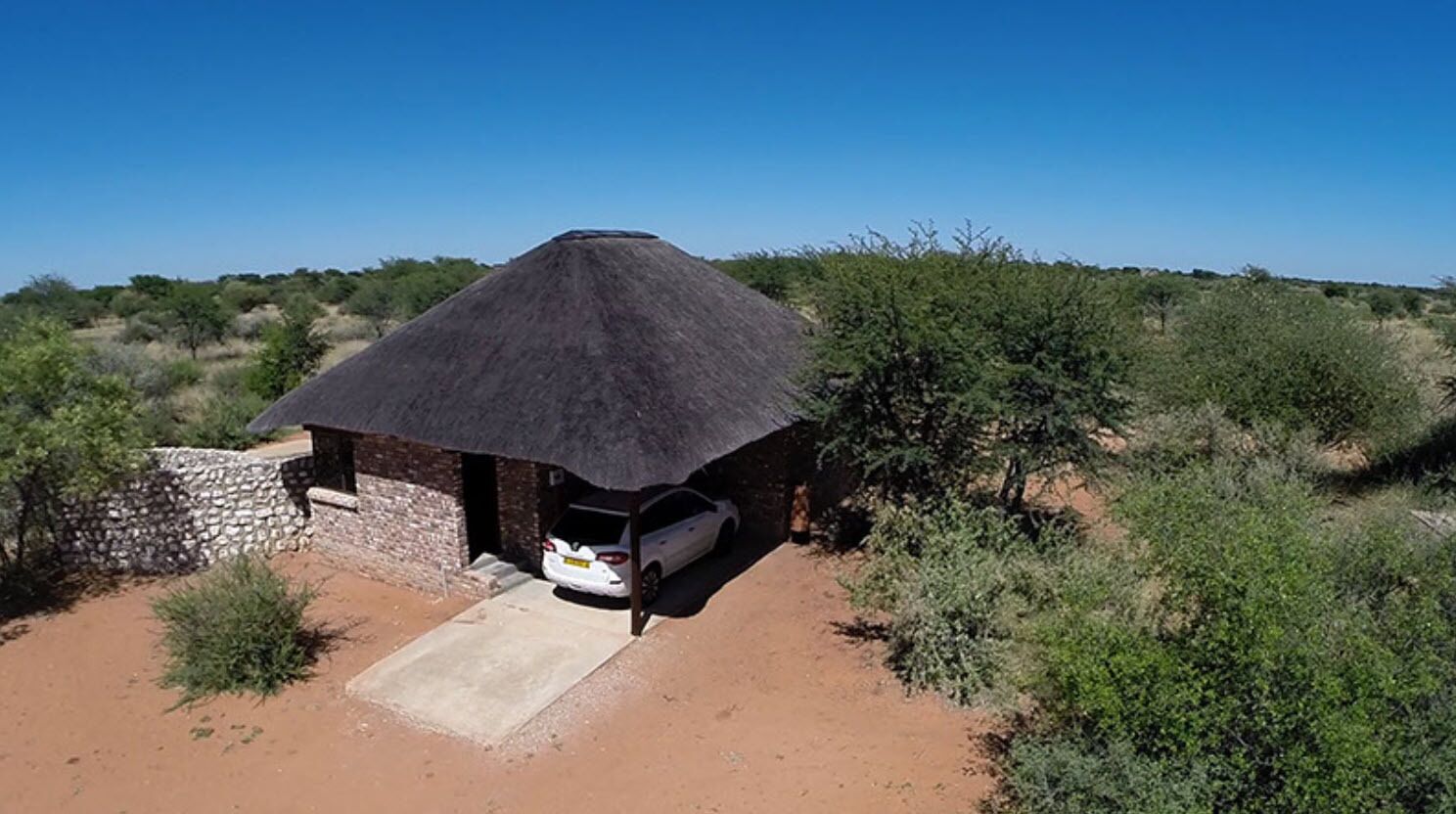 A thatched-roof hut with a car parked outside, surrounded by sparse vegetation.