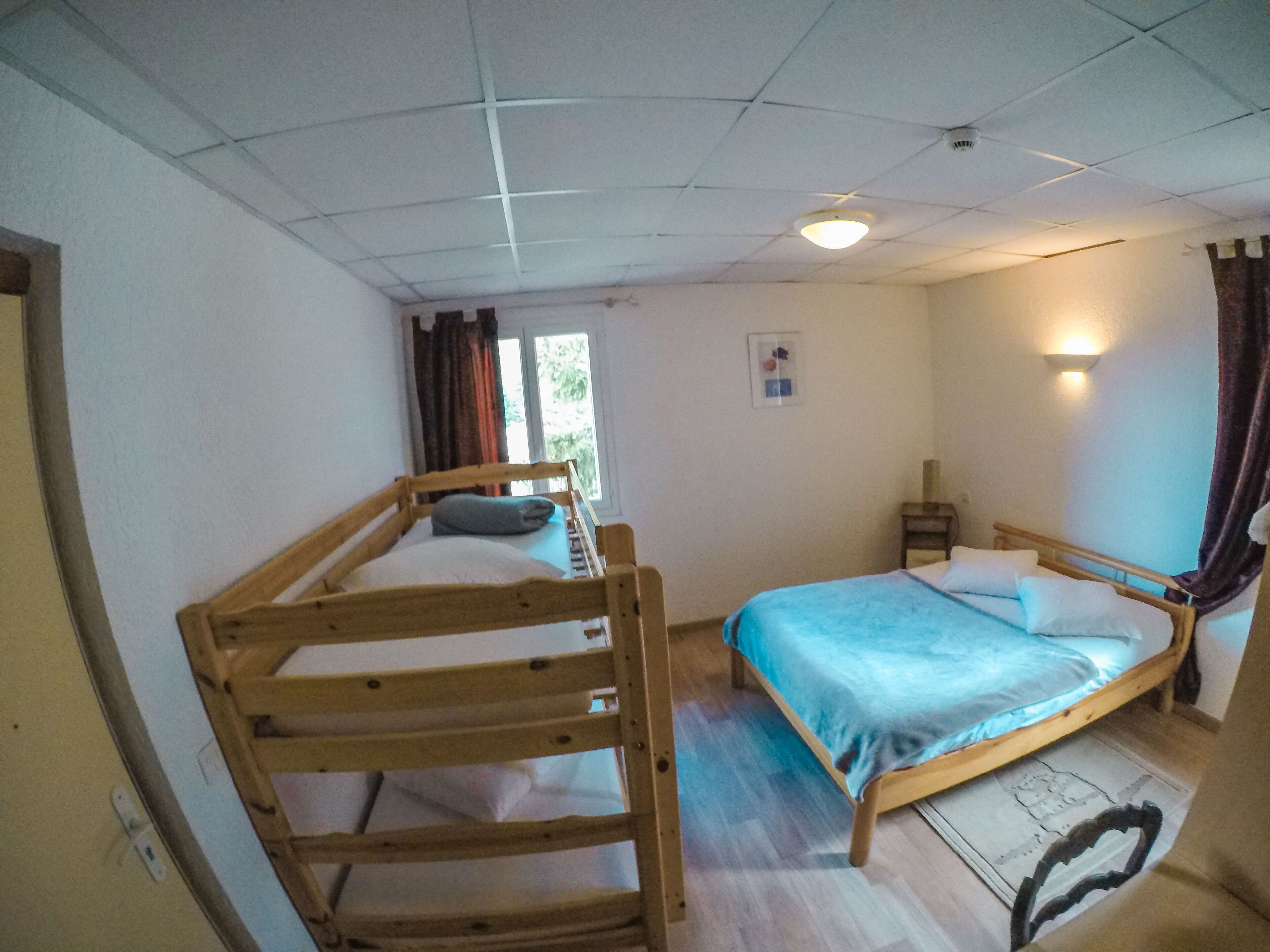 A room with a bunk bed, a single bed, a small bedside table, a wall-mounted light, and a window with curtains.