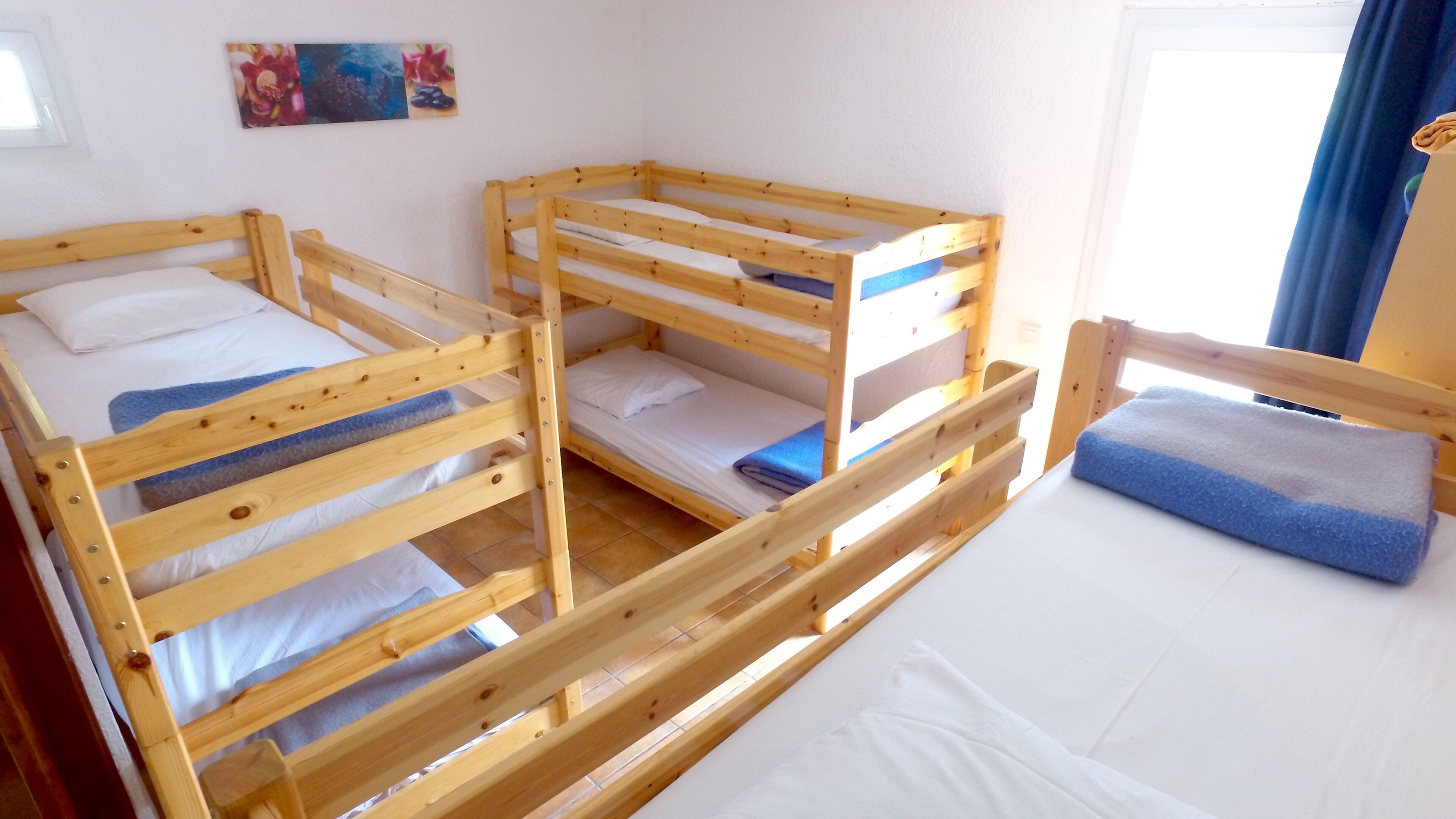Ein Zimmer mit drei Holzkohlschränken, jeweils mit Kissen und Decke. Ein Fenster mit blauen Vorhängen und ein Gemälde an der Wand.