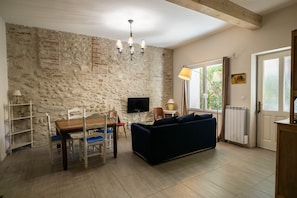A living room with a stone wall, a wooden dining table with chairs, a blue sofa, a TV, and a window with curtains.