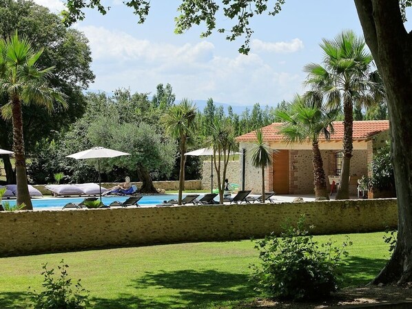 A poolside area with lounge chairs and umbrellas, surrounded by palm trees and a building with a red-tiled roof. - Domaine Castell de Blés - Gîte Vermeille (Saint-Genis-des-Fontaines)
