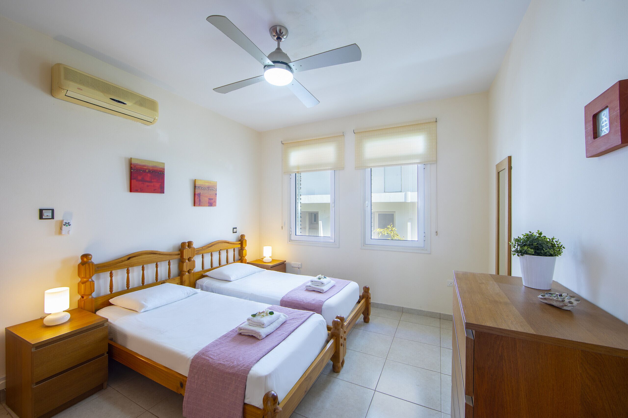 Une chambre avec deux lits, un ventilateur de plafond, un climatiseur, une table de chevet avec une lampe et une commode agrémentée d’une plante.