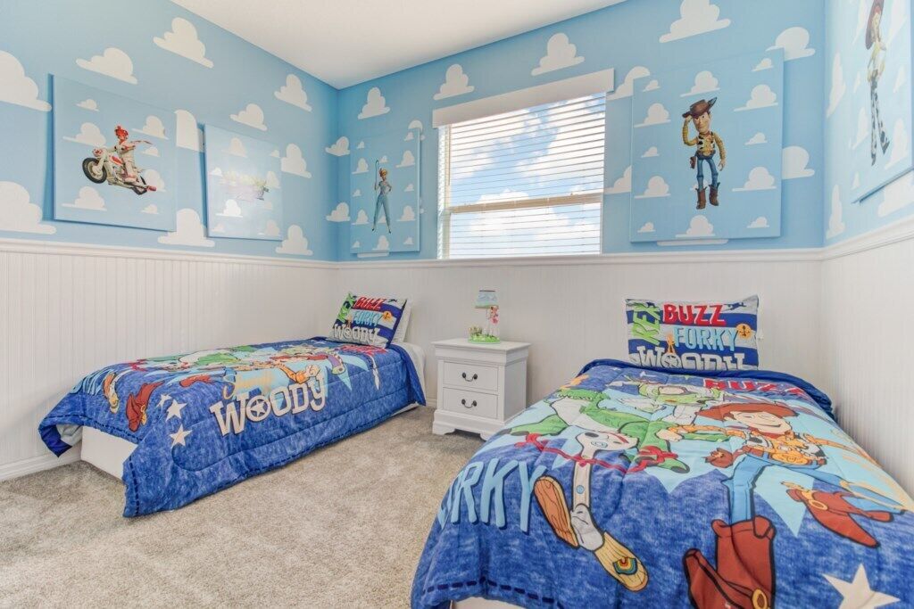 A bedroom with two beds, each with a blue bedspread featuring cartoon characters. The walls are painted light blue with white clouds, and there are framed pictures on the walls.