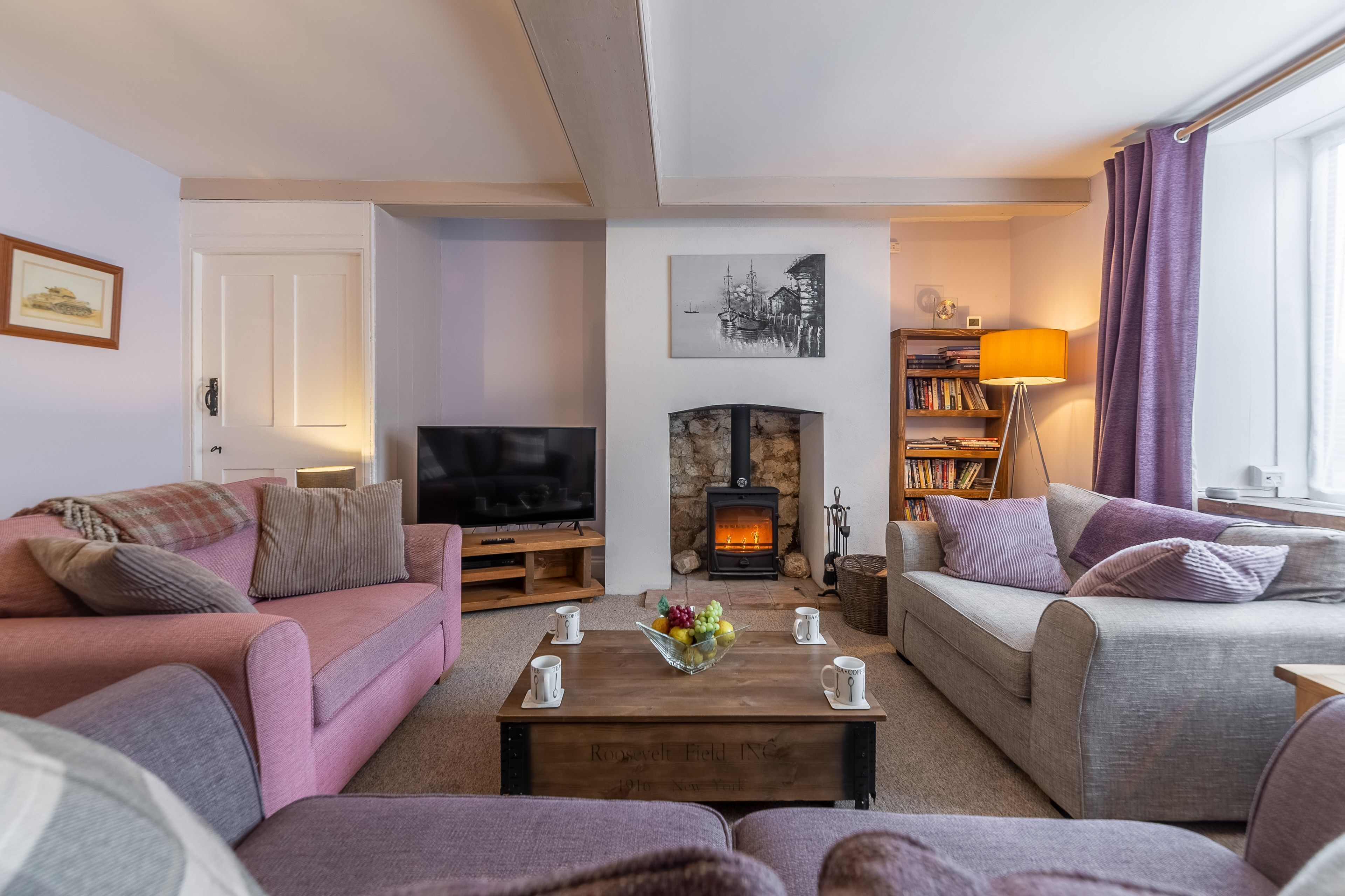A cozy living room with a fireplace, sofas, a coffee table with a fruit bowl, and a bookshelf.